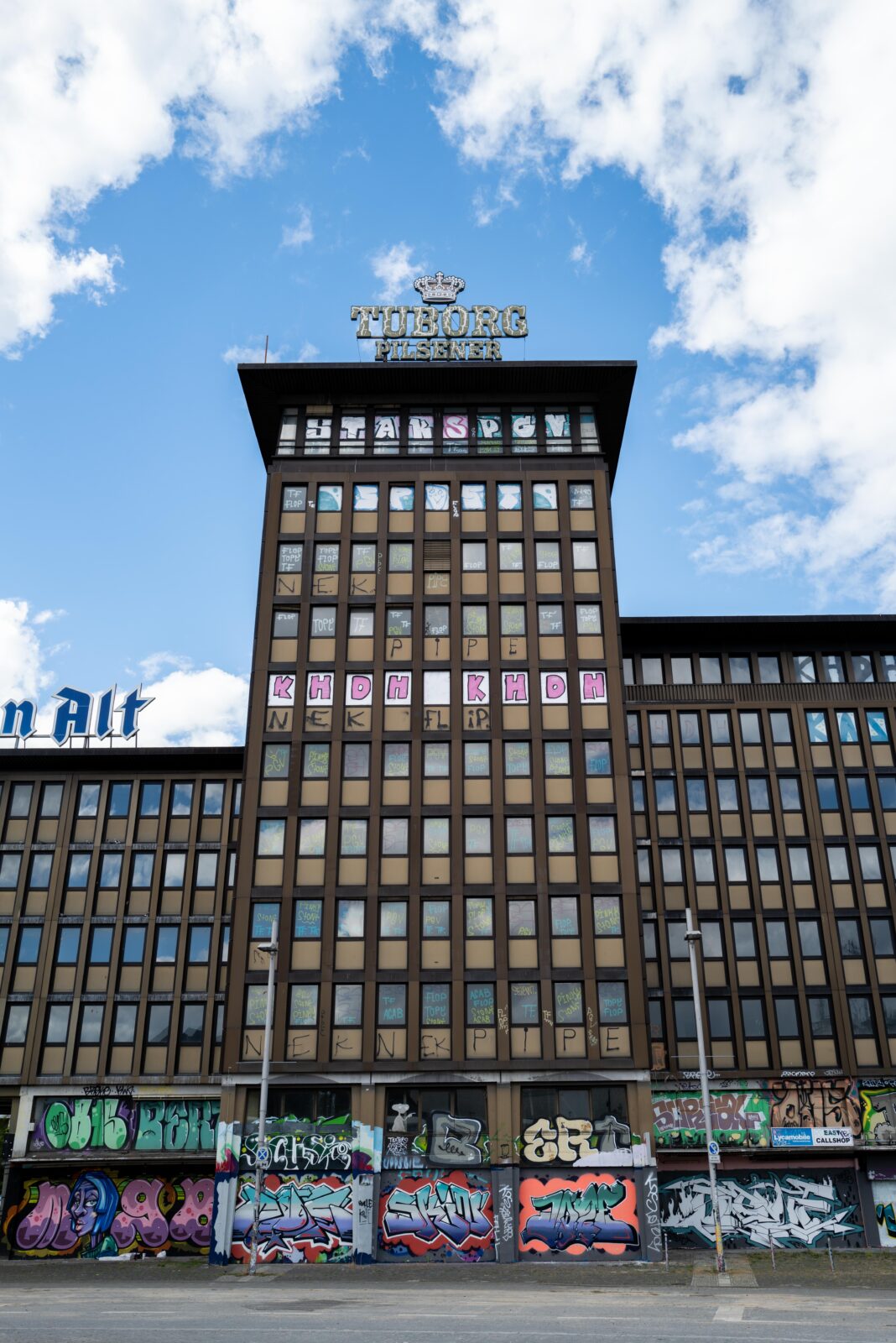 Das leerstehende Hochhaus Haus Westland in Mönchengladbach, mit großen Schildern ‚Tuborg Pilsener‘ und ‚Schumacher Alt‘ auf dem Dach, zahlreichen Graffiti an den unteren Fassadenbereichen und bemalten Fenstern in den oberen Etagen. Der Himmel ist blau mit einigen Wolken.